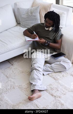 Uomo adulto africano che scrive con una penna in un quaderno con bordi rossi su un tappeto, tablet a fianco, con cuffie Foto Stock