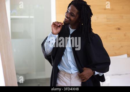 Uomo afroamericano che sistema la toga accademica e controlla il riflesso nello specchio della camera da letto Foto Stock
