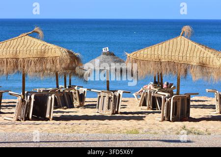L'immagine sembra mostrare una scena di spiaggia sabbiosa con ombrelloni di paglia e sedie a sdraio impilate lungo la costa, probabilmente nella regione dell'Algarve di Foto Stock