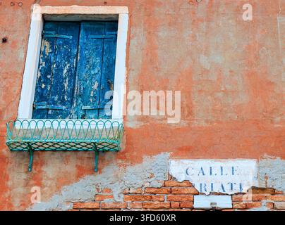 Persiane blu di legno consumate e un piccolo balcone in ferro battuto si trovano in alto su un muro di terracotta sbiadito, con l'intonaco che si sfalda e rivela il vecchio mattone. Foto Stock