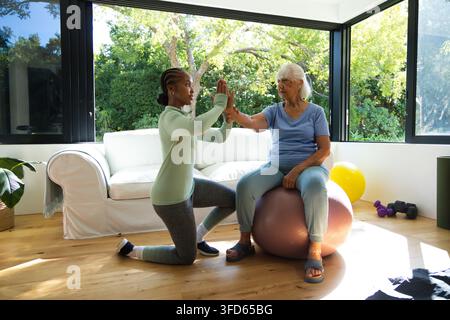 Badante afroamericana inginocchiata e guida un'anziana su un pallone da ginnastica a casa con leggings Foto Stock