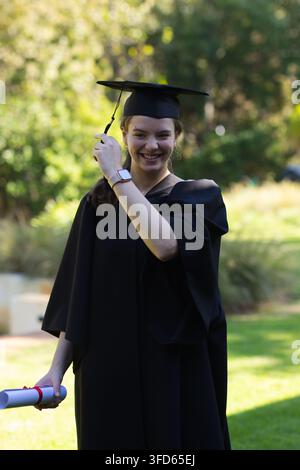 Donna laureata in posa sul prato, indossa abito nero e toga, tiene il diploma, tocca la nappina, smartwatch Foto Stock