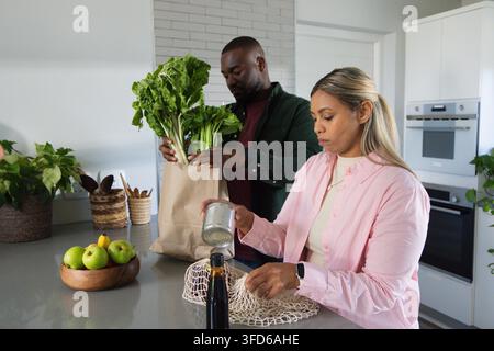 Coppia afroamericana che disimballa la spesa nella cucina di casa, maneggiando un sacchetto di carta, una lattina di metallo, olio d'oliva Foto Stock