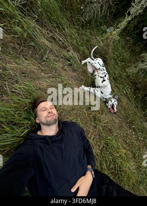 Selfie ad angolo alto di un giovane uomo barbuto in felpa con cappuccio nera che si rilassa sull'erba selvatica con gli occhi chiusi accanto a un felice cane dalmata sdraiato sulla schiena sulla pancia Foto Stock