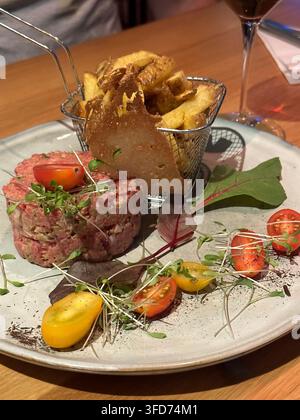 Tartara di manzo gourmet servita con patatine fritte croccanti e pomodorini freschi. Tartara di manzo gourmet ben presentata su un piatto di ceramica con rossi a metà a Foto Stock