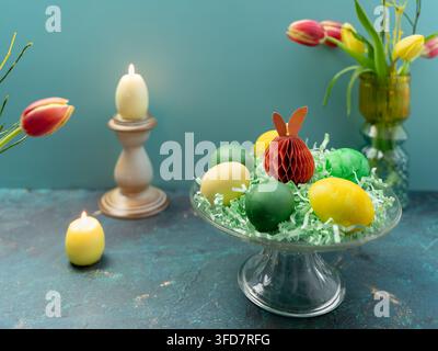 Bellissimo arrangiamento pasquale con una candela, uova decorative colorate, tulipani freschi e ornamenti a forma di coniglio, stilizzati come una natura morta festiva primaverile. Foto Stock