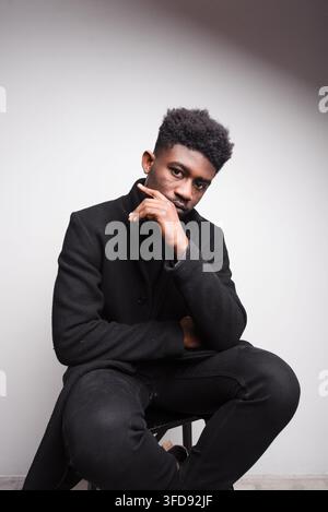 Un giovane uomo con la pelle scura e i capelli ricci è seduto in un ambiente da studio, con uno sguardo contemplativo e la mano sul mento Foto Stock
