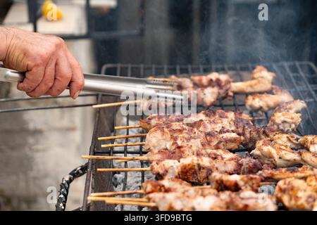Prima piano di un'azione che mostra la mano di una persona che usa delle pinze metalliche per girare con cura degli spiedini di souvlaki di maiale greci tradizionali grigliati su carbone fumante Foto Stock