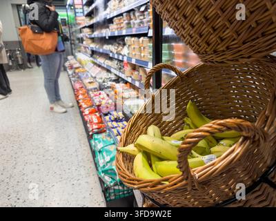 Le banane riempiono un cesto di vimini all'ingresso di un corridoio di prodotti alimentari di un supermercato. Una cliente con una borsa a tracolla arancione sfoglia gli scaffali. Londra, Regno Unito Foto Stock