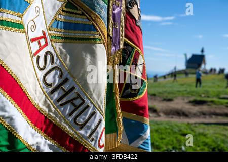 Aschau, Germania. 30 agosto 2026. Una bandiera con l'iscrizione "Aschau" si trova davanti alla Cappella commemorativa Maria, Regina della Pace sul Kampenwand prima dell'inizio della messa di montagna. Questo fine settimana segna anche la prima volta che la croce di vetta più alta delle Alpi bavaresi·che è stata convertita all'illuminazione a LED·sarà illuminata. Credit: Peter Kneffel/dpa/Alamy Live News Foto Stock