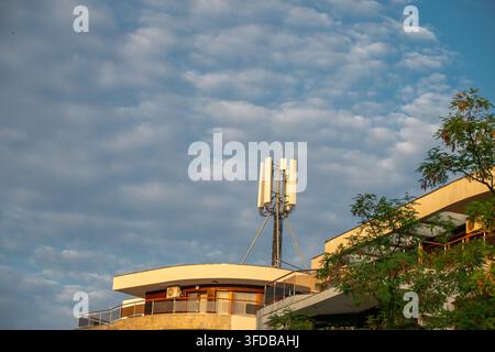 Un'antenna di cella telecomunicazioni sul tetto è montata su un edificio moderno contro un cielo blu nuvoloso al tramonto. Foto Stock