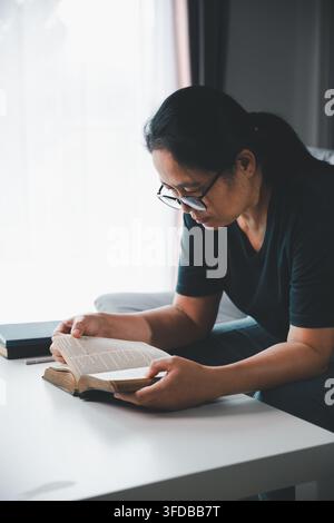 Donna di mezza età pensierosa con occhiali che legge una Bibbia aperta a casa, simboleggiando la devozione cristiana, lo studio delle Scritture, la fede, la saggezza, a Foto Stock