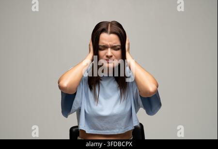 Giovane donna che si copre le orecchie con le mani, prova mal di testa o stress da rumore forte, soffre di sintomi di emicrania Foto Stock