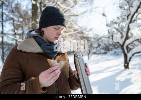 Giovane uomo che beve tè caldo e mangia un panino nel parco invernale Foto Stock