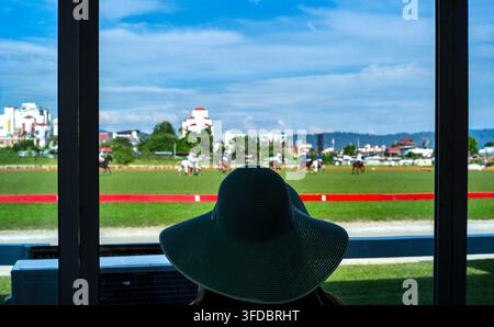 Una fotografia incorniciata da interno verso esterno che cattura la silhouette di uno spettatore con un cappello di paglia a tesa larga visto di spalle, mentre osserva una partita di polo Foto Stock