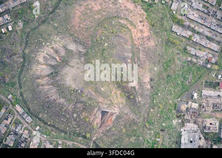 Cumulo di scorie a forma di cuore e cumulo di scorie minerarie danneggiato circondati da strutture industriali in rovina e edifici devastati, vista aerea dall'alto Foto Stock
