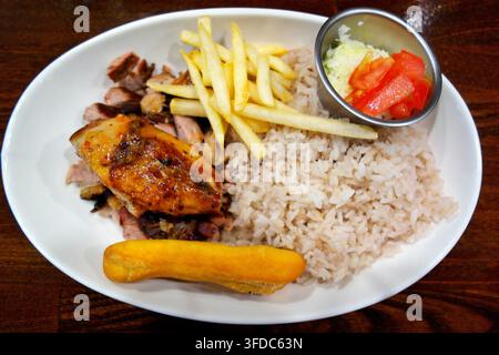 Delizioso pollo e maiale jerk caraibico servito con riso condito, patatine fritte, impasto fritto festivo e un contorno di insalata di pomodori freschi in una ciotola bianca Foto Stock