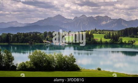 Una vasta veduta del Forggensee rivela una campagna verde, un piccolo insediamento e una catena montuosa alpina frastagliata sotto nuvole stratificate. Alberi e arbusti Foto Stock