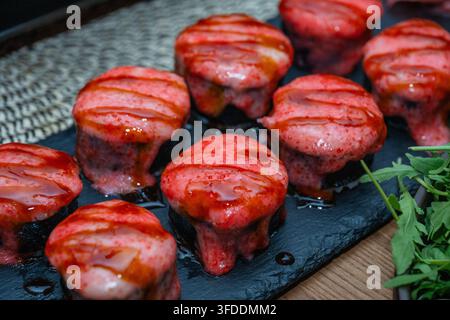 Una vista dall'alto mostra più rotoli di sushi cotti al forno guarniti con una salsa cremosa rosa e una glassa di soia dolce disposti su un piatto di ardesia scura accanto a verdure fresche Foto Stock