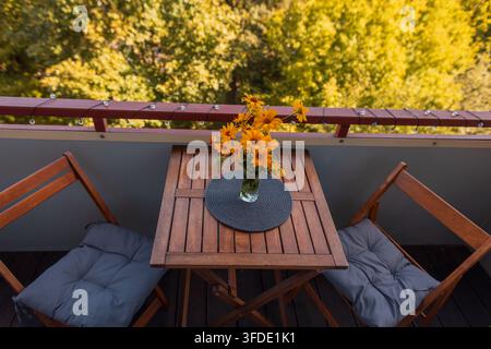 Ambientazione accogliente sul balcone con un tavolo di legno e fiori gialli brillanti in una vaso, due sedie pieghevoli con cuscini e una vista panoramica su un albero autunnale Foto Stock