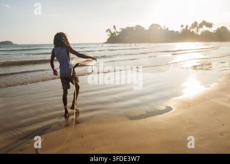 Uomo nero con capelli lunghi che tiene una tavola da surf bianca, cammina sulla sabbia bagnata verso le onde dell'oceano alla bellissima spiaggia di Dewata nello Sri Lanka durante la calda ora dorata Foto Stock