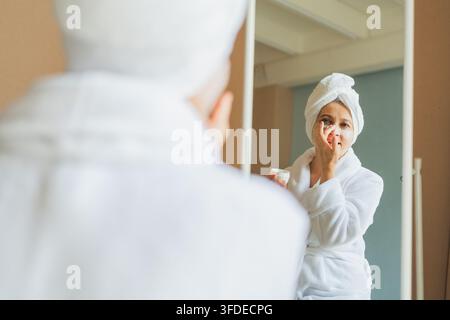 Matura donna anziana adulta che guarda lo specchio applicando una crema viso nutriente anti età facendo routine di bellezza quotidiana. Donna anziana con crema maschera bianca sulla pelle ottiene trattamento viso cura della pelle invecchiamento della pelle Foto Stock