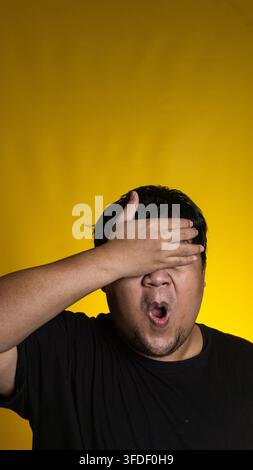 Un giovane asiatico scioccato si copre gli occhi con una mano mentre tiene la bocca spalancata contro uno sfondo giallo brillante. L'individuo espressivo Foto Stock