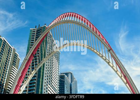 Prima piano ad angolazione bassa di un arco decorativo in acciaio bianco e rosso davanti a grattacieli moderni in vetro sotto un cielo azzurro e soleggiato Foto Stock