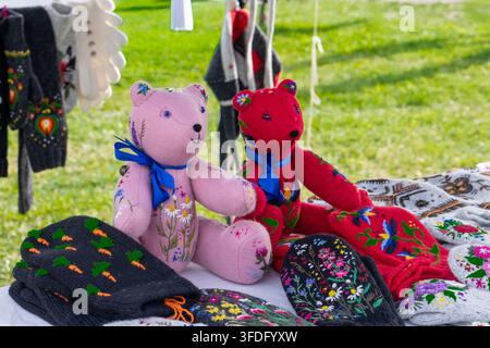Orsi di peluche rosa e rossi fatti a mano decorati con ricami floreali seduti su un tavolo in un mercato artigianale all'aperto. Orsi giocattolo in tessuto morbido con nastri blu Foto Stock