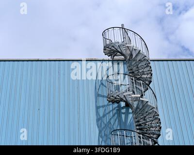 Scatto architettonico di una moderna scala a chiocciola in acciaio zincato montata sul lato di una parete di edificio in metallo blu chiaro. Presenta una geometria chiara Foto Stock