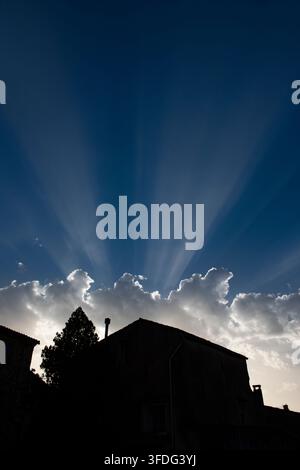 Silhouette di una casa e di un albero sotto un cielo drammatico con raggi di sole che emergono da dietro le nuvole Foto Stock
