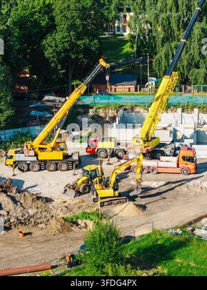 Vista aerea di un cantiere edile con gru mobili gialle, un escavatore e un camion parcheggiato su un lotto di terra circondato da alberi verdi rigogliosi a Riga Foto Stock
