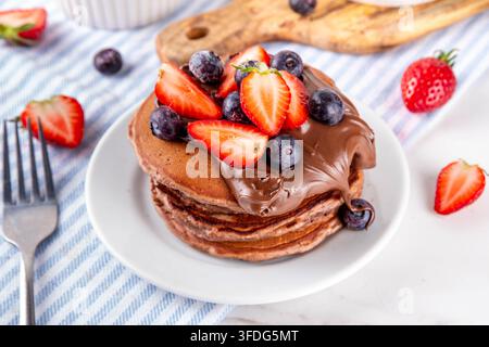 Pila di pancake al cioccolato con una ricca crema di pasta al cioccolato e frutti di bosco freschi - fragole, mirtilli. Colazione dolce e gustosa Foto Stock