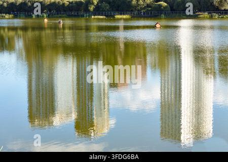 Gli edifici moderni si riflettono nella calma acqua del lago, circondato da una vegetazione rigogliosa, mentre delle casette per anatre in legno galleggiano in superficie. Foto Stock