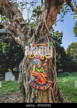 14 ott 2025 Dehradun India. Statua colorata della divinità indù Radha Krishna appesa a un grande tronco d’albero in un giardino all’aperto verde con cielo blu e natura Foto Stock