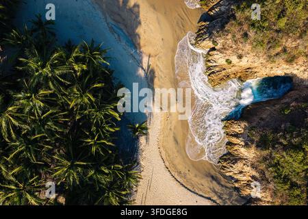 Vista aerea della costa a Maruata, con una spiaggia sabbiosa, palme rigogliose, scogliere rocciose e onde turchesi che si infrangono contro la riva a Mar Foto Stock