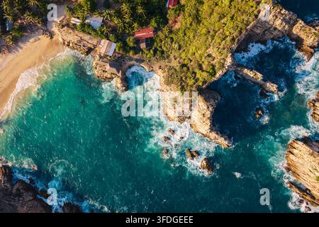 Vista aerea della spiaggia di Maruata con acque turchesi, scogliere rocciose frastagliate, palme lussureggianti e piccoli edifici costieri a Maruata, Michoacan, Messico Foto Stock