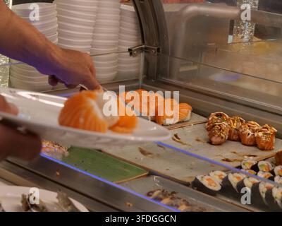 Vista laterale di un diner che usa delle pinze metalliche per prendere sushi nigiri al salmone fresco e metterlo su un piatto bianco in una stazione di buffet all'interno di un Ja Foto Stock