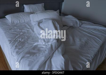 Letto d'albergo disfatto con lenzuola bianche sgualcite, cuscini e copriletto in una stanza degli ospiti fioca dopo il sonno. Alloggio di viaggio quotidiano, disordine mattutino, comfort, res Foto Stock