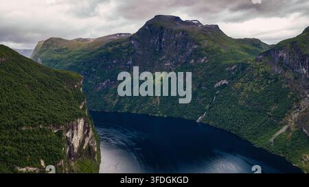Prospettiva aerea di un lago alpino profondo circondato da pareti rocciose verticali, fitta vegetazione forestale verde e un picco montuoso lontano. Foto Stock
