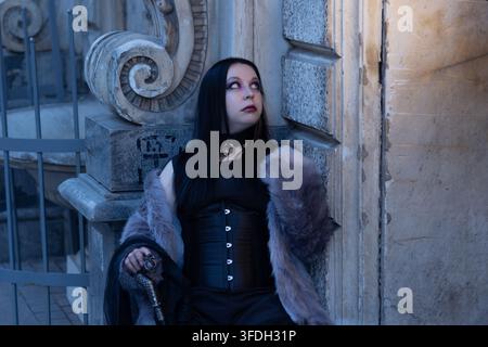 Donna vampira gotica in pelliccia e corsetto che riposa su un muro di pietra, immagine di personaggio fantasy oscuro. Pelle pallida e trucco scuro alla luce fredda della sera. Foto Stock