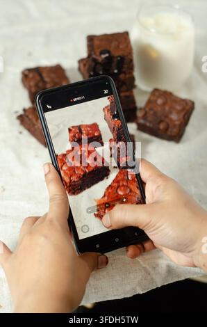 Mani che usano uno smartphone per fotografare brownies al cioccolato fatti in casa per il food blogging e i social media. Foto Stock