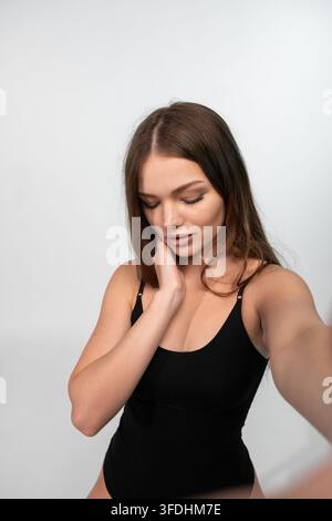 Giovane donna che si scatta un selfie, si tocca il collo, mostra bellezza naturale e amore per sé stessa su sfondo bianco Foto Stock