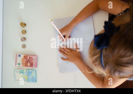 Bambino che scrive in un quaderno accanto a banconote e monete in euro Foto Stock