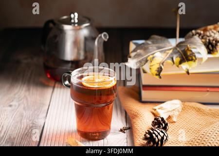 Tè autunnale appena preparato in una tazza di vetro accanto a libri impilati su un tavolo di legno rustico, vista laterale, stile cupo e atmosferico, spazio per il testo Foto Stock
