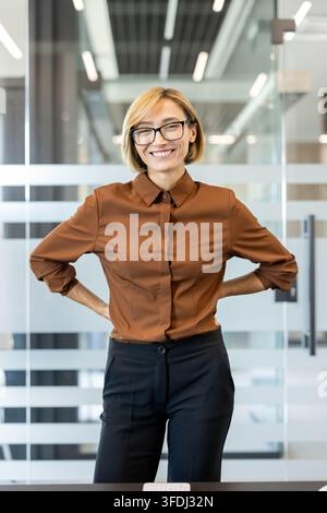 Donna d'affari sicura di sé che sorride, indossa occhiali e una camicia marrone, in piedi con le mani sui fianchi, esprimendo successo e leadership in un ambiente d'ufficio contemporaneo Foto Stock