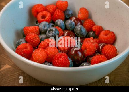 Prima vista di lamponi, mirtilli e ciliegie disposti in una ciotola blu e bianca maculata su un tavolo di legno con frutta mista colorata Foto Stock
