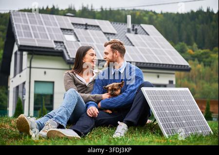 Coppia seduta sull'erba con un piccolo cane vicino a casa moderna con pannelli solari, guardarsi l'un l'altro. Sistema fotovoltaico sul tetto, energia rinnovabile, stile di vita ecologico Foto Stock