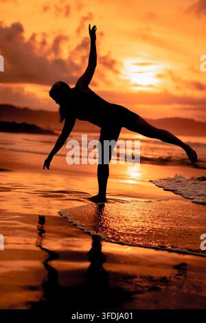 Una donna crea una drammatica posa di equilibrio su una gamba sola sulla spiaggia, incorniciata da brillanti colori del tramonto e riflessi specchiati. Foto Stock