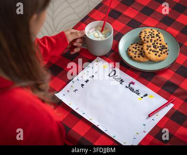 Bambino che tiene una tazza di bevanda calda con marshmallow accanto a un piatto di biscotti al cioccolato su una tovaglia a quadri rossa in pelle di bufalo Foto Stock